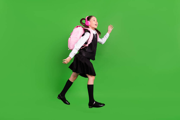 Foto di affascinante eccitata ragazza della scuola indossare cuffie uniforme zaino cercando spazio vuoto saltando isolato sfondo di colore verde - Foto, immagini