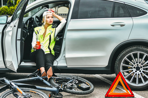 girl in emergency vest in the car drinks beer. the bike lies on the asphalt - Foto, Imagem