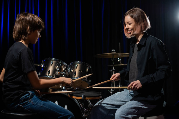 Young caucasian woman teaches a boy to play the drums in the studio on a black background. Music school student - Fotoğraf, Görsel