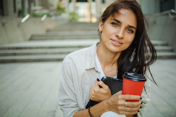 Schöne Geschäftsfrau wird mit Kaffee zu Fuß in der Nähe von Bürogebäuden arbeiten. Porträt einer erfolgreichen Geschäftsfrau, die auf dem Weg zur Arbeit eine Tasse Heißgetränk in der Hand hält. - Foto, Bild