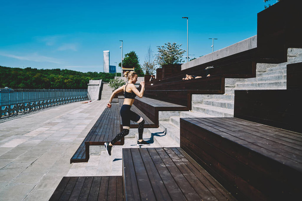 Side view of fast focused female in sport clothes doing sport exercise running up stairs at promenade under bright sun - Φωτογραφία, εικόνα