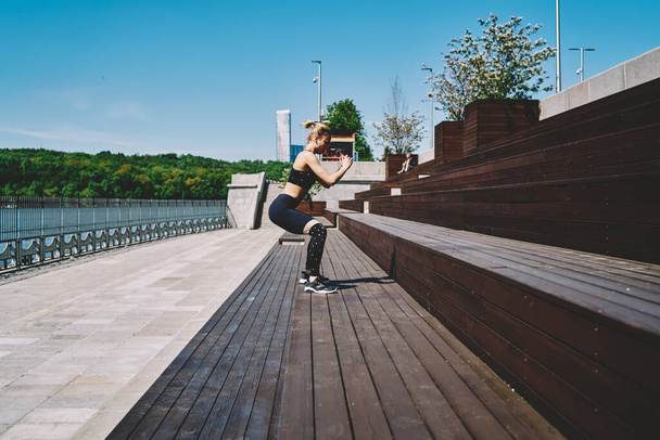 Vue latérale de la jeune femme active en vêtements de sport faisant des exercices de fitness sautant sur les escaliers au bord de l'eau sous un soleil éclatant - Photo, image