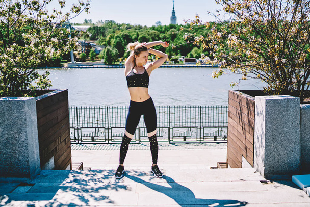 Fit female gymnast in black fashionable active wear standing while doing warm up with hands and looking away at quay on sunny day  - Zdjęcie, obraz