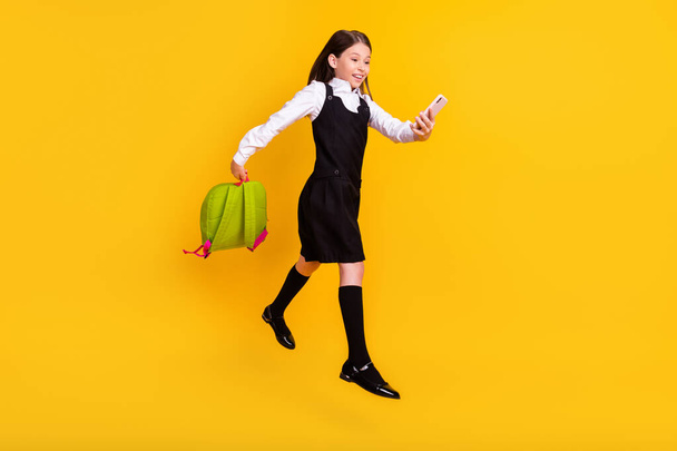 Full length body size photo schoolgirl jumping with rucksack using smartphone isolated vibrant yellow color background - Photo, Image