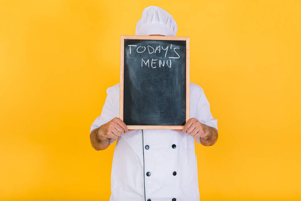 Шеф-повар в белой фуражке и куртке закрывает лицо доской с надписью "Сегодняшнее меню" на желтом фоне. - Фото, изображение