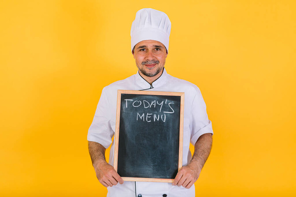 Chef cuisinier portant chapeau de cuisine blanc et veste tenant un tableau noir qui dit "Menu du jour", sur fond jaune - Photo, image