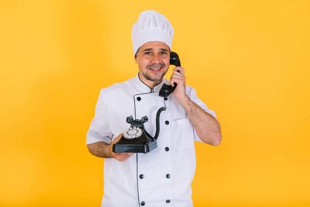 Chef cuisinier vêtu d'une casquette et d'une veste de cuisine blanche parle sur un téléphone rétro, sur fond jaune - Photo, image