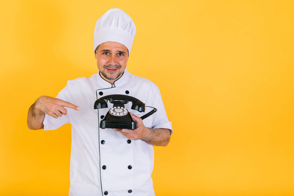 Chef cuisinier portant casquette de cuisine blanche et veste tenant et pointant vers un téléphone rétro, sur fond jaune - Photo, image