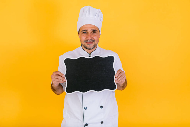 Chef kok draagt witte keukenkap en jas met een zwart krijtbord, op gele achtergrond - Foto, afbeelding