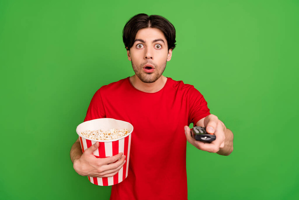 Photo of speechless stunned guy hold popcorn box remote control wear red t-shirt isolated green color background - Fotoğraf, Görsel