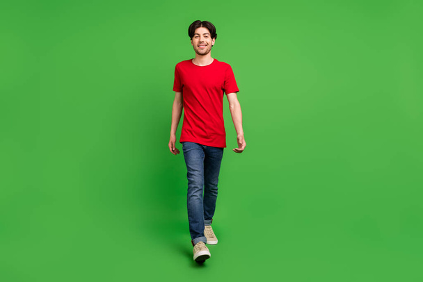 Photo of relaxed positive guy walk enjoy street stroll wear red t-shirt jeans shoes isolated green color background - Photo, Image