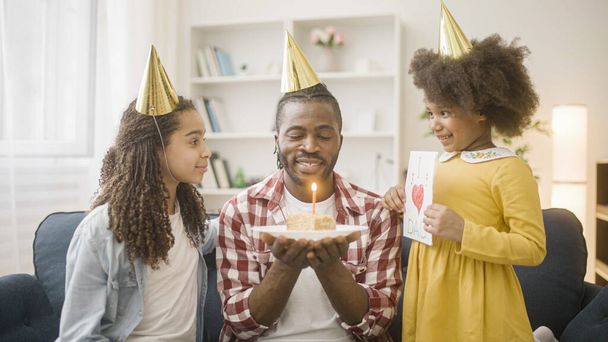 Joyful dad in party hat blowing out birthday cake candle, celebrating with daughters - Photo, image