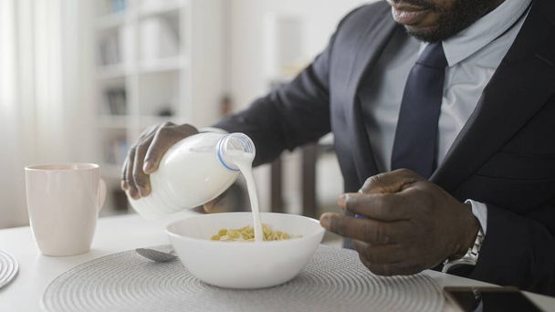 Black businessman in suit pouring milk into cereal bowl, morning routine - Foto, afbeelding
