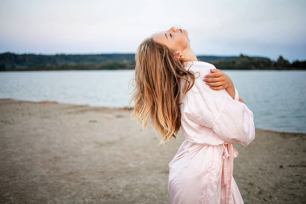 Romantyczna blondynka w różowym szlafroku na plaży o zachodzie słońca. - Zdjęcie, obraz