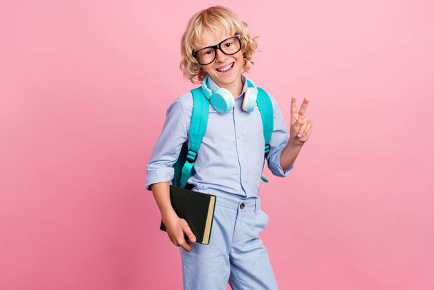 Photo of pretty attractive schoolboy wear spectacles rucksack headphones v-sign holding book isolated pink color background - Fotoğraf, Görsel