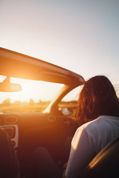Rearview of an attractive brunette woman riding in the convertible car. Concept of summer adventures and new experiences. Strong sunlight during warm and long summer evenings. - Photo, Image