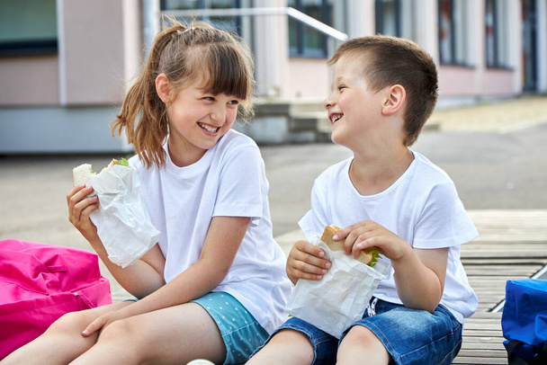 Schoolchildren boy and girl laugh and eat their lunch, snack, breakfast in the school yard. Food for children in educational institutions, kindergartens. - Photo, Image