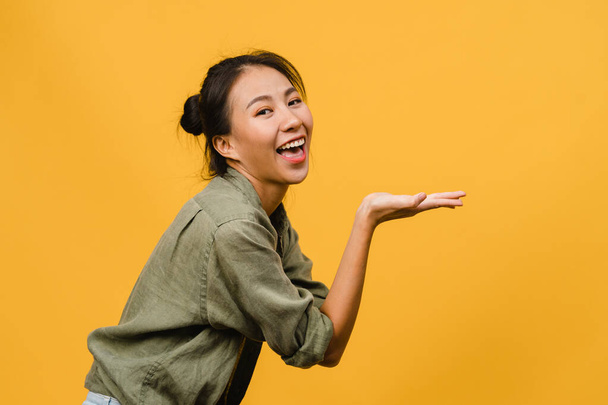 Portrait einer jungen Asiatin, die mit fröhlichem Gesichtsausdruck lächelt, etwas Erstaunliches in einem leeren Raum in lässigem Tuch zeigt und isoliert in die Kamera auf gelbem Hintergrund blickt. Gesichtsausdruck. - Foto, Bild