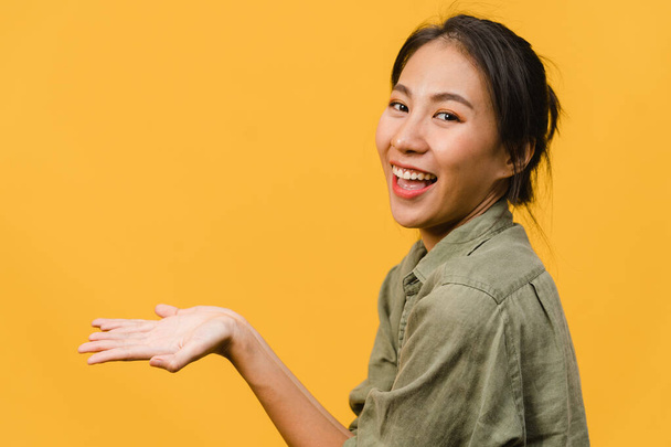 Portrét mladé asijské dámy usmívající se s veselým výrazem, ukazuje něco úžasného na prázdný prostor v ležérní látky a při pohledu na kameru izolované přes žluté pozadí. Koncept výrazu obličeje. - Fotografie, Obrázek