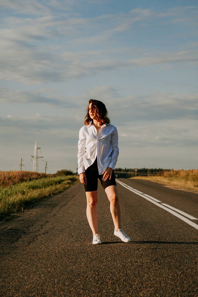 A red-haired woman in a white shirt walks along the road among the fields - Foto, Bild