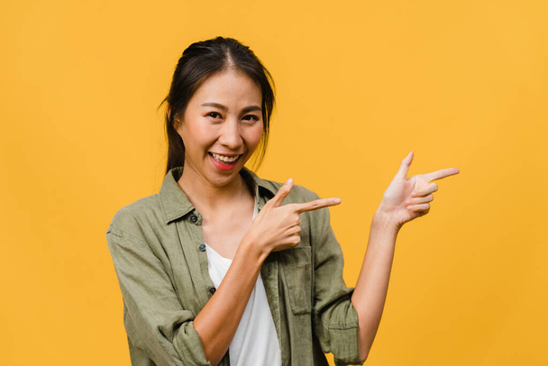 Portret van jonge Aziatische dame glimlachend met vrolijke uitdrukking, toont iets verbazingwekkends op lege ruimte in casual doek en kijkend naar camera geïsoleerd over gele achtergrond. Gezichtsuitdrukking. - Foto, afbeelding