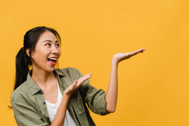 Portrait of young Asian lady smiling with cheerful expression, shows something amazing at blank space in casual clothing and standing isolated over yellow background. Facial expression concept. - Photo, Image