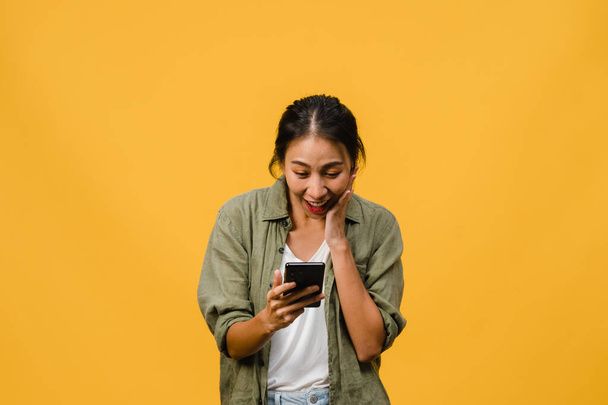 Young Asia lady using phone with positive expression, smiles broadly, dressed in casual clothing feeling happiness and stand isolated on yellow background. Happy adorable glad woman rejoices success. - Valokuva, kuva
