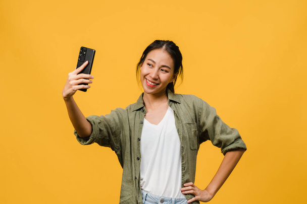 Smiling adorable Asian female making selfie photo on smart phone with positive expression in casual clothing and stand isolated on yellow background. Happy adorable glad woman rejoices success. - Foto, Imagem