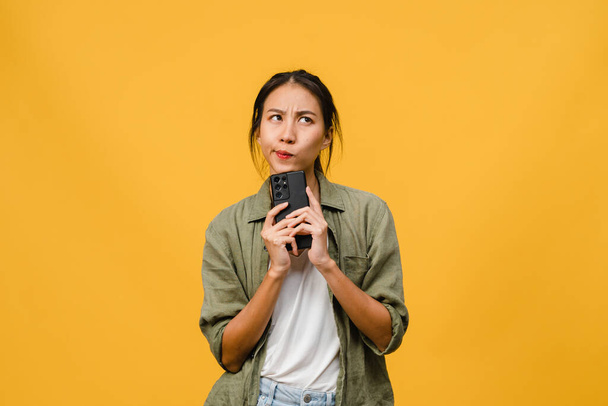 Thinking dreaming young Asia lady using phone with positive expression, dressed in casual cloth feeling happiness and stand isolated on yellow background. Happy adorable glad woman rejoices success. - Photo, Image