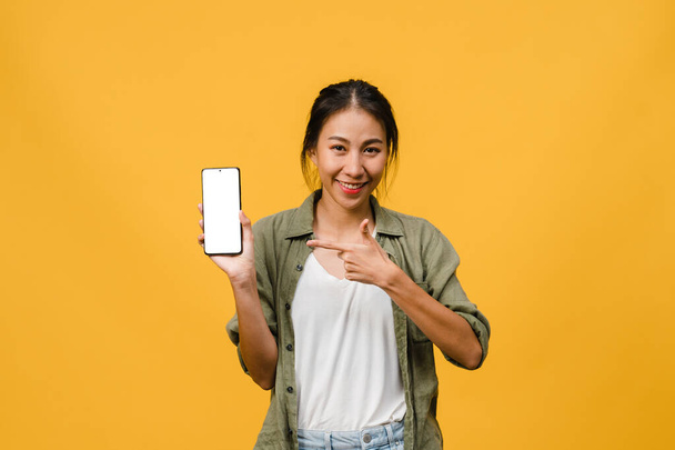 Młoda Azjatka pokazuje pusty ekran smartfona z pozytywnym wyrazem twarzy, uśmiecha się szeroko, ubrana w luźną odzież, odczuwając szczęście na żółtym tle. Telefon komórkowy z białym ekranem w kobiecej dłoni. - Zdjęcie, obraz