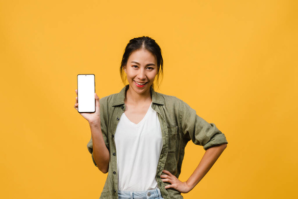 Молода азіатка показує порожній екран смартфона з позитивним виразом, посміхається широко, одягнена в повсякденний одяг, відчуваючи щастя на жовтому тлі. Мобільний телефон з білим екраном у жіночих руках.. - Фото, зображення