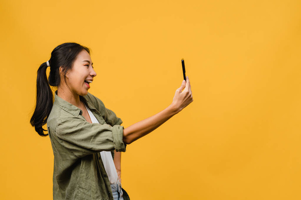 Smiling adorable Asian female making selfie photo on smart phone with positive expression in casual clothing and stand isolated on yellow background. Happy adorable glad woman rejoices success. - Photo, image