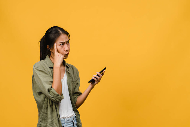 Thinking dreaming young Asia lady using phone with positive expression, dressed in casual cloth feeling happiness and stand isolated on yellow background. Happy adorable glad woman rejoices success. - Foto, immagini