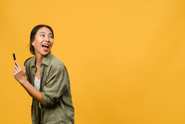 Young Asia lady using phone with positive expression, smiles broadly, dressed in casual clothing feeling happiness and stand isolated on yellow background. Happy adorable glad woman rejoices success. - Photo, Image