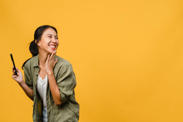 Young Asia lady using phone with positive expression, smiles broadly, dressed in casual clothing feeling happiness and stand isolated on yellow background. Happy adorable glad woman rejoices success. - Photo, image