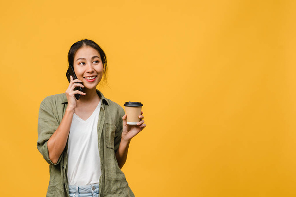 Young Asia lady talk by phone and hold coffee cup with positive expression, smile broadly, dressed in casual cloth feeling happiness and stand isolated on yellow background. Facial expression concept. - Photo, image
