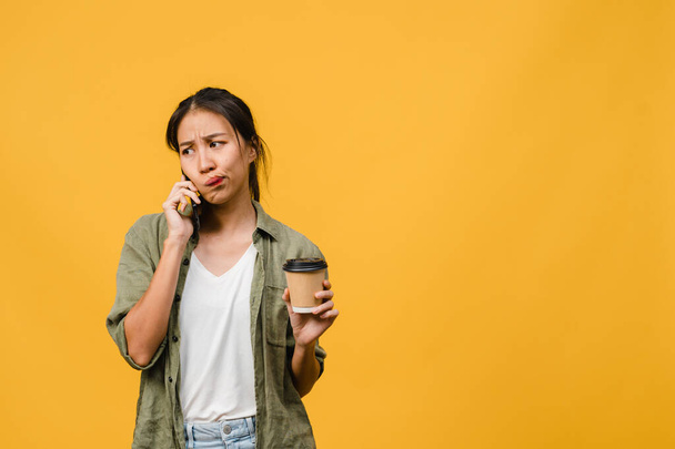 Genç Asyalı bayan telefonla konuşuyor ve elinde negatif bir ifade olan kahve bardağını tutuyor. Heyecanlı çığlıklar atıyor, duygusal olarak ağlıyor ve sarı arka planda tek başına duruyor. Yüz ifadesi kavramı. - Fotoğraf, Görsel