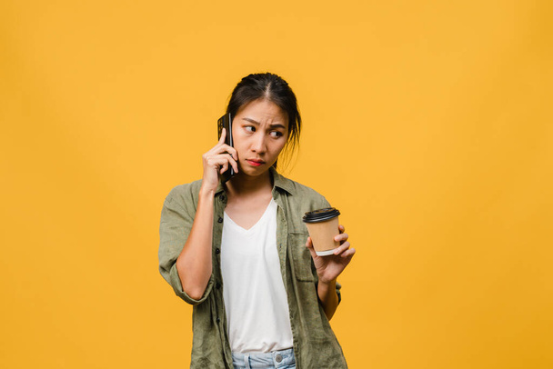 Young Asia lady talk by phone and hold coffee cup with negative expression, excited screaming, cry emotional angry in casual cloth and stand isolated on yellow background. Facial expression concept. - Φωτογραφία, εικόνα