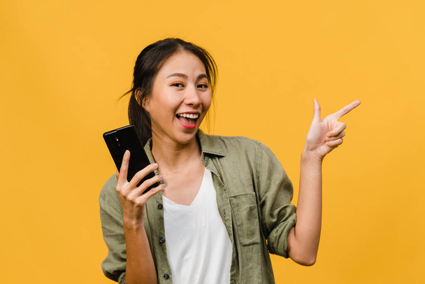 Young Asian lady using mobile phone with cheerful expression, shows something amazing at blank space in casual cloth and looking at camera isolated over yellow background. Facial expression concept. - Photo, Image