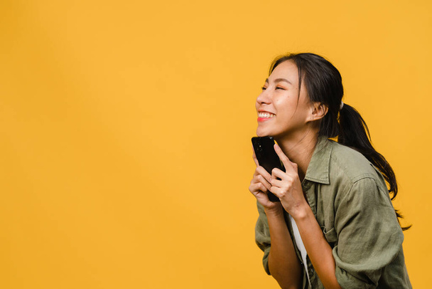 Nuori Aasian nainen käyttää puhelimen positiivinen ilme, hymyilee laajasti, pukeutunut rento vaatteet tunne onnea ja seistä eristetty keltaisella taustalla. Onnellinen ihana iloinen nainen iloitsee menestyksestä. - Valokuva, kuva