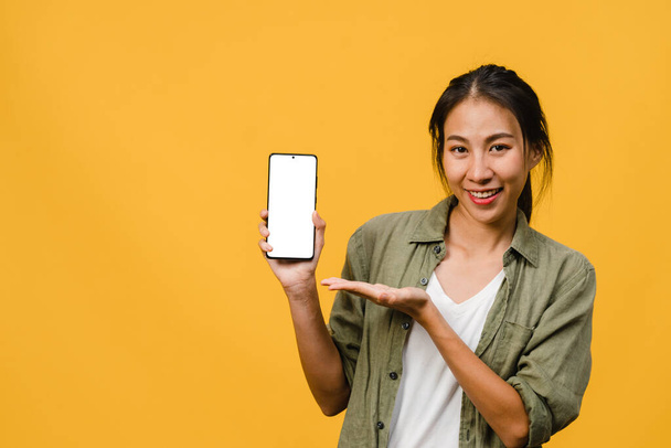 Jonge Asia dame tonen lege smartphone scherm met positieve expressie, glimlacht breed, gekleed in casual kleding gevoel geluk op gele achtergrond. Mobiele telefoon met wit scherm in vrouwelijke hand. - Foto, afbeelding