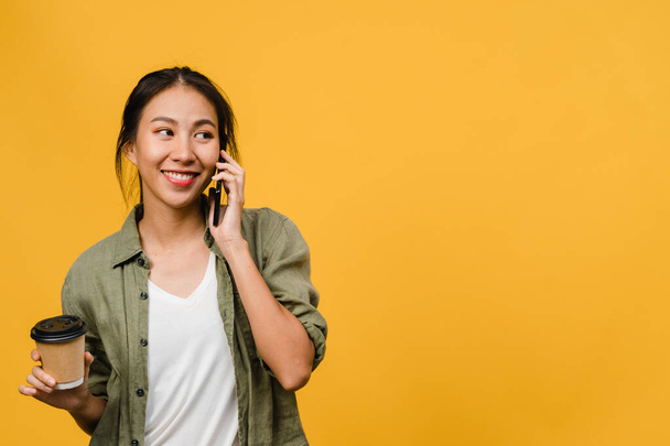 Young Asia lady talk by phone and hold coffee cup with positive expression, smile broadly, dressed in casual cloth feeling happiness and stand isolated on yellow background. Facial expression concept. - Valokuva, kuva
