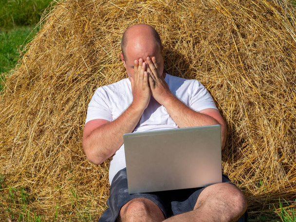 Mies istuu heinäsuovassa läppärin kanssa ja peittää silmänsä käsillään. Etätyö, verkkokonferenssi maaseudulta - Valokuva, kuva