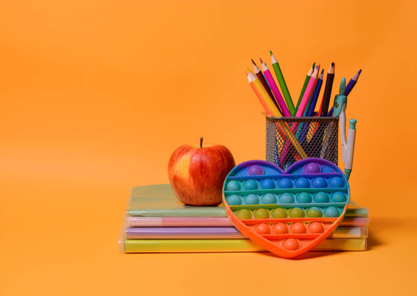 Schoolbenodigdheden: notitieboekjes, boeken, kleurpotloden, een appel, pop it - Nieuwe populaire kleurrijke siliconen zintuiglijke trendy speelgoed antistress op een oranje achtergrond. Terug naar school. Kopieerruimte. - Foto, afbeelding