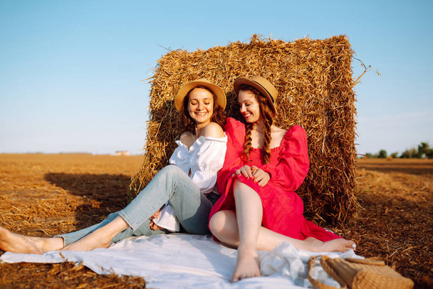 Two smiling young woman resting near haystack. Fashion concept. Nature, vacation, relax and lifestyle. - Fotografie, Obrázek