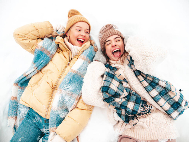 Two young beautiful smiling hipster female in trendy warm clothes and scarfs.Carefree women posing in the street in park. Positive pure models lying in snow. Enjoying winter moments. Xmas concept - Valokuva, kuva