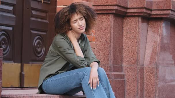 Mujer afroamericana cansada joven se sienta en las escaleras masajeando el cuello frotando los músculos tensos después de un largo viaje a pie por la ciudad. Postura incorrecta, estilo de vida sedentario, necesidad de ejercicios o concepto de tratamiento - Imágenes, Vídeo