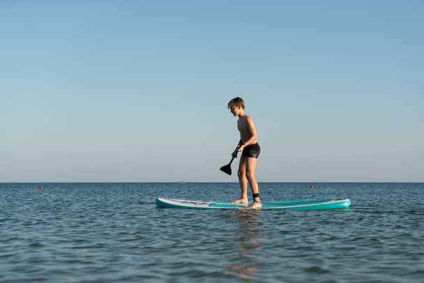 Un garçon de 12 ans apprend à se tenir debout sur une planche SUP dans la mer près du rivage - Photo, image