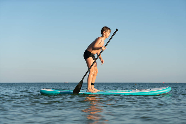 12-летний мальчик учится стоять на доске SUP в море недалеко от берега - Фото, изображение