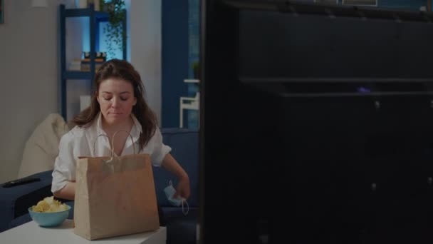 Kobieta z maską na twarzy siedzi na kanapie z dostawą żywności - Materiał filmowy, wideo